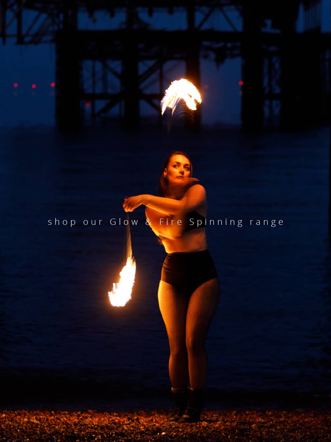 Fire poi being spun by a person standing on a pebble beach in front of a burnt out pier at night